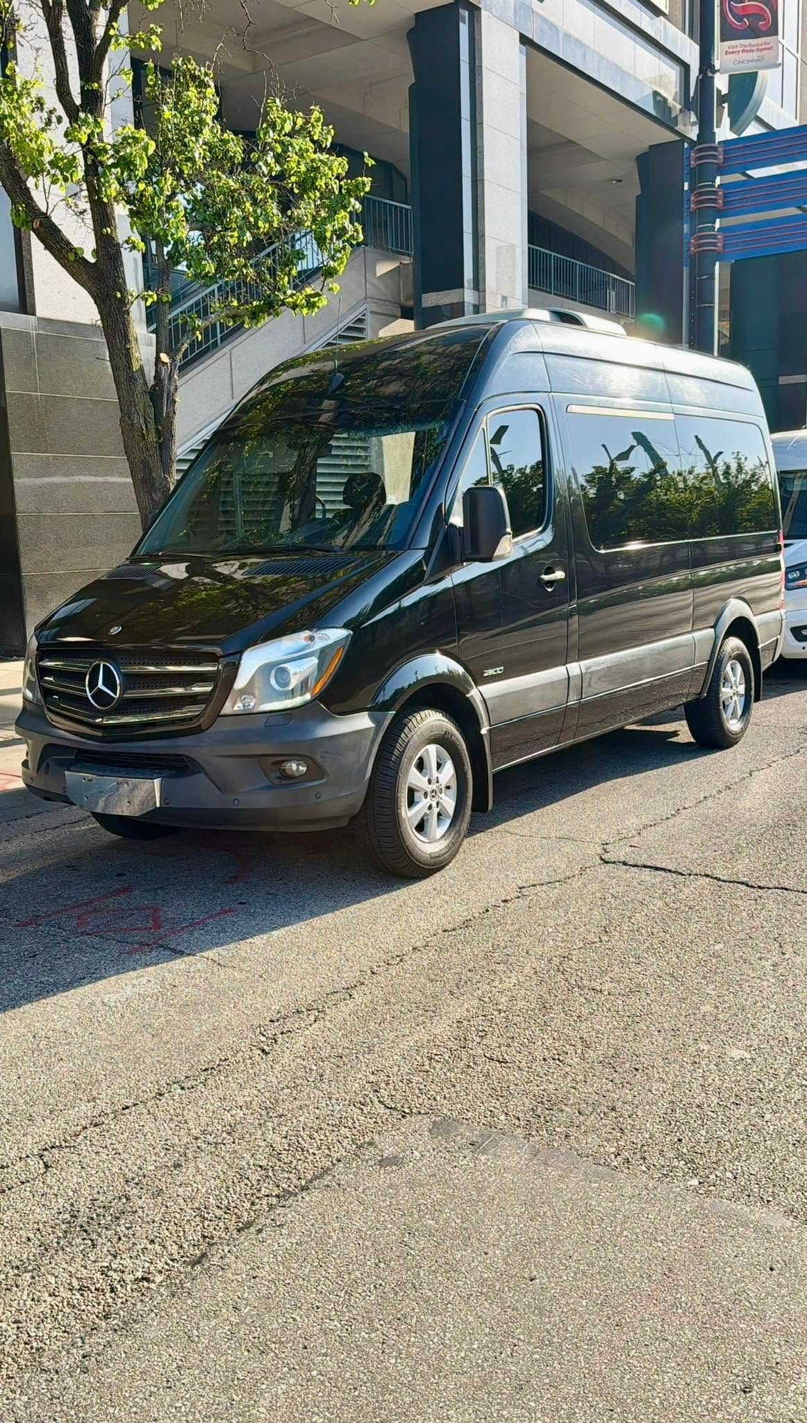Black van parked on a sunlit cobblestone street beside a building and tree