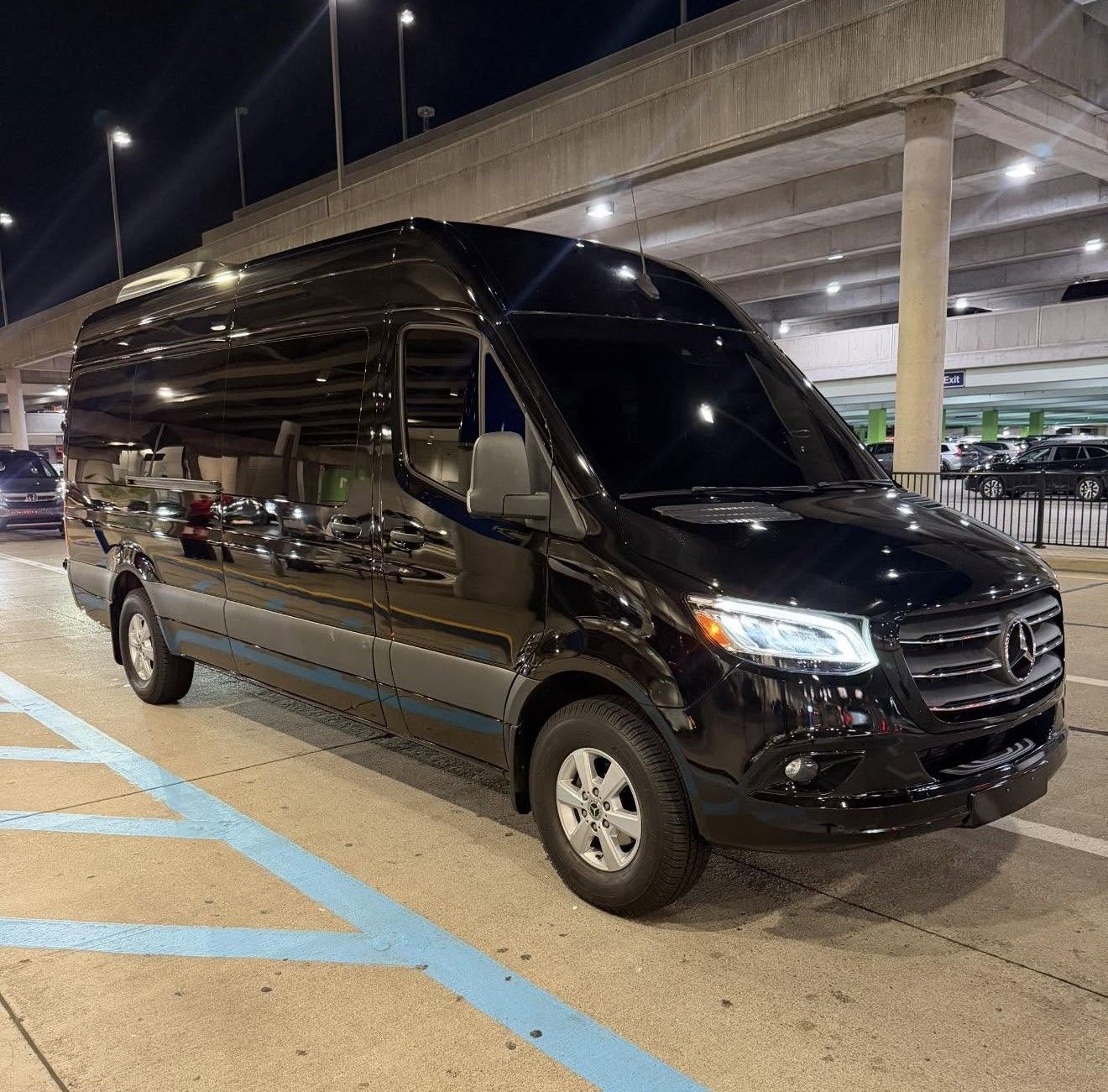 Black passenger van parked at night under a highway overpass, with blue-striped pavement nearby