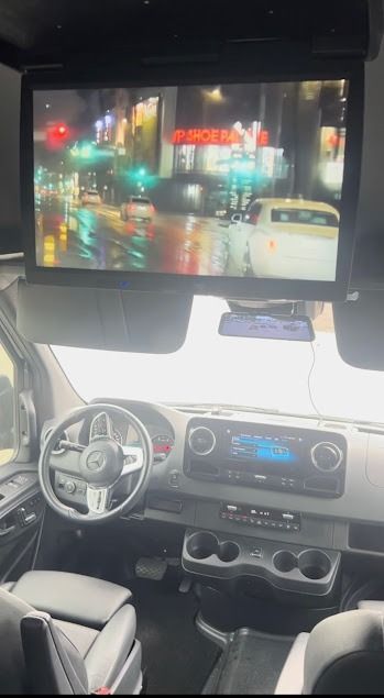 Car interior with dashboard and steering wheel, viewed from the back seat, with a large screen showing a rainy street at night.