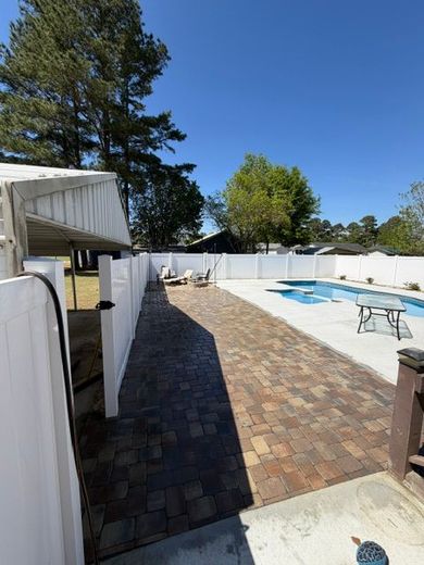Backyard patio with brick walkway, white fence, trees, and an above-ground pool under blue sky