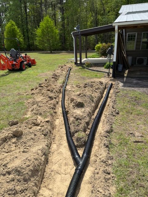 Black corrugated drainage pipes laid in a trench beside a house, with an orange tractor in the yard.