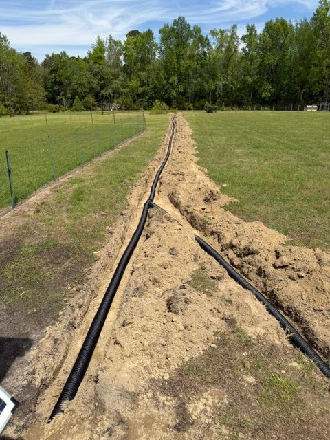 Black irrigation hoses run through a sandy trench across a grassy field toward trees.