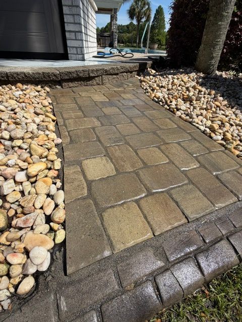 Stone paver walkway leading to a pool area, bordered by decorative river rocks and palm trees