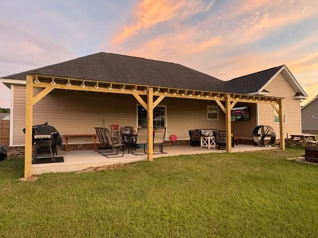 Covered patio with seating and grill beside a house, viewed across a grassy yard at sunset.