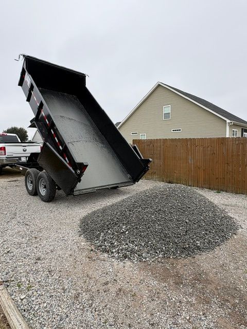 Dump truck unloading gray gravel on a driveway beside a fenced suburban house.
