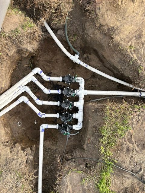 Trench with white PVC irrigation pipes and a black manifold valve assembly in bare soil.