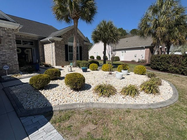 Front yard with white rock landscaping, trimmed shrubs, and palm trees beside a brick house and driveway.
