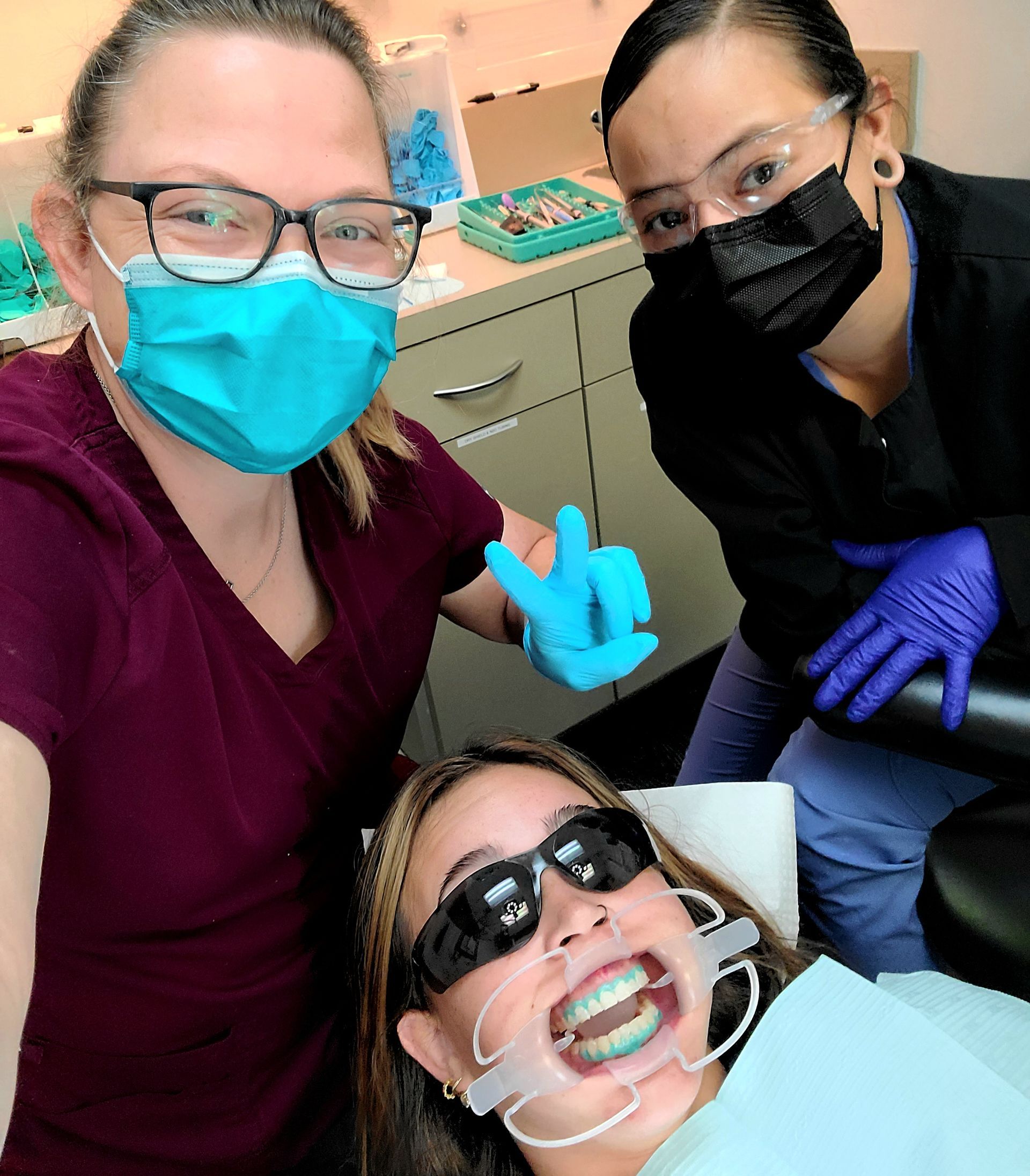 Dentist and two masked assistants smiling around a patient in a dental chair, wearing gloves and safety gear.