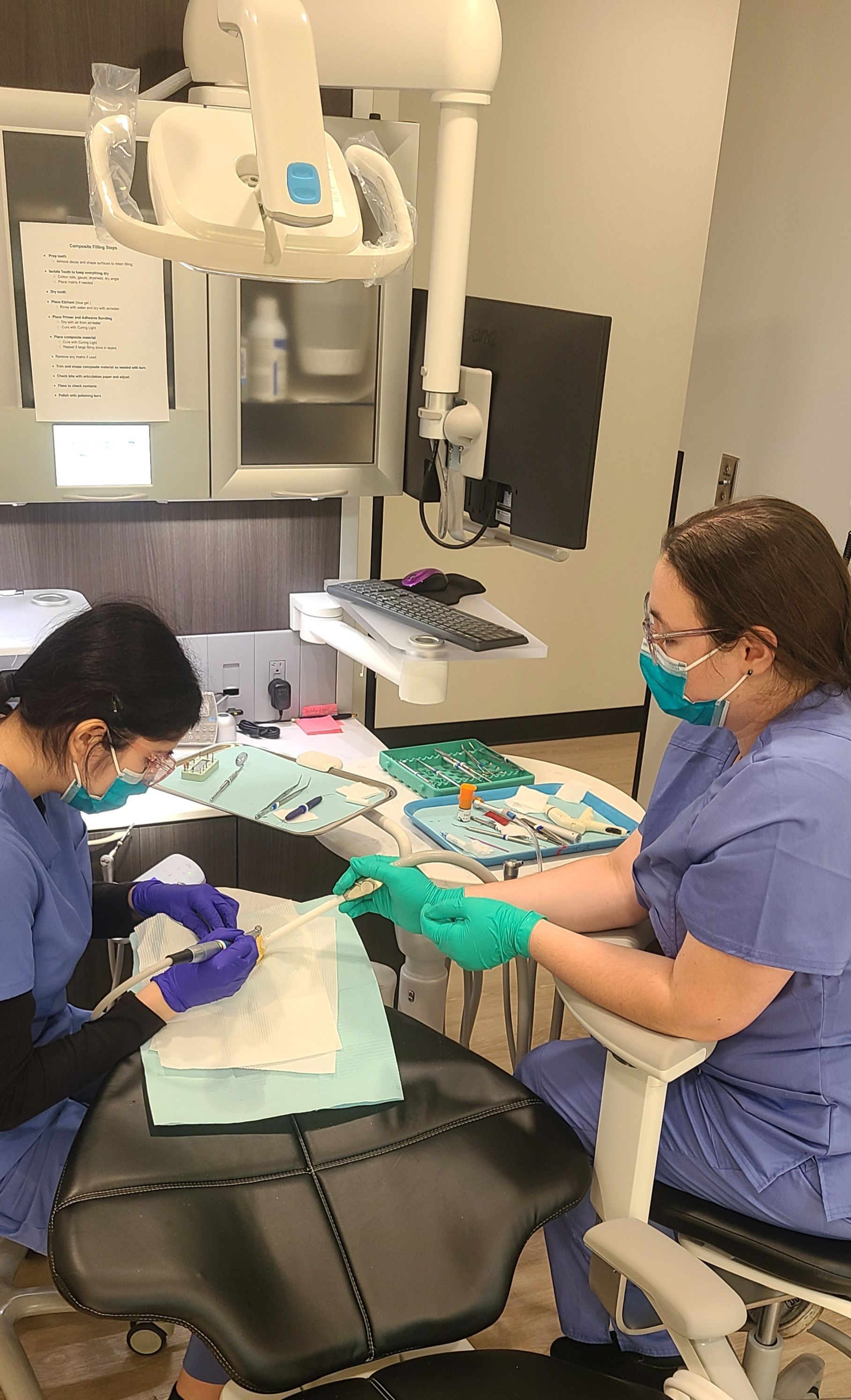 Dental assisting students  practicing on patient in a clinic, with one clinician adjusting tools under an overhead exam light.