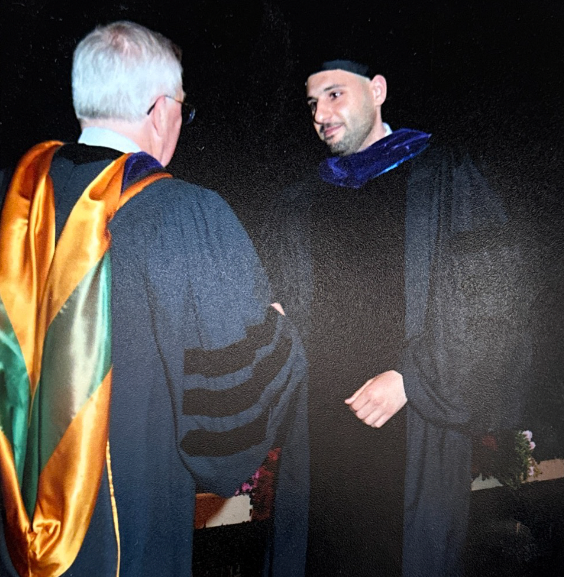 Man in graduation gown shakes hands with another person in academic regalia.