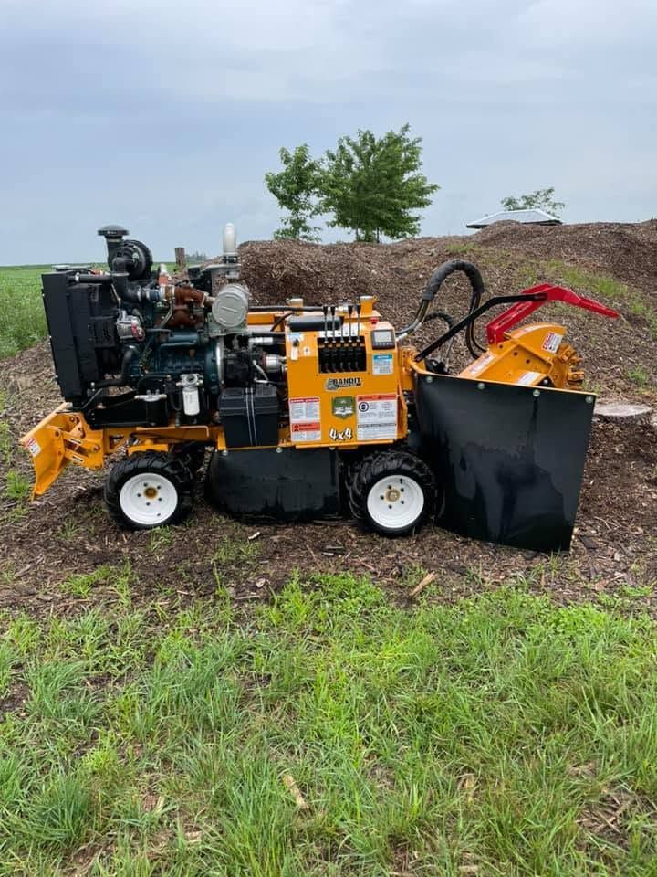 Orange wood chipper on a grassy field beside a dirt mound and tree under an overcast sky