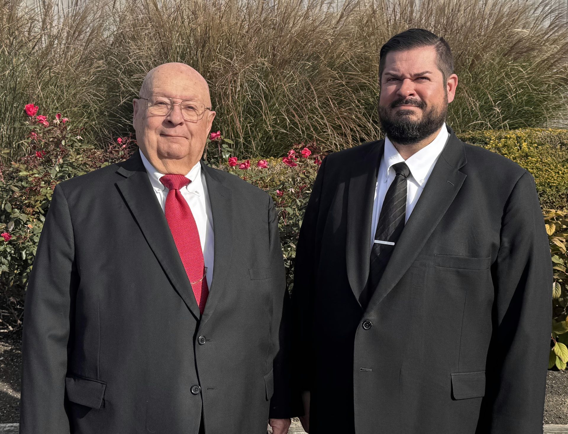 Two men in black suits standing outdoors in front of bushes, one wearing a red tie and smiling.