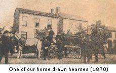 Horse-drawn hearses in front of a building, with people standing nearby, circa 1870.