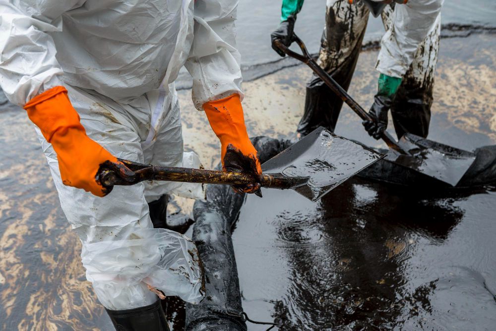 Workers in hazmat suits and orange gloves shovel oil from a contaminated surface.