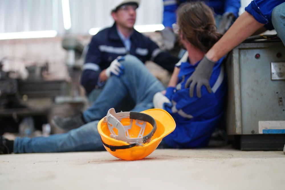 Worker injured on a factory floor, colleagues assisting. Yellow hard hat on the floor.