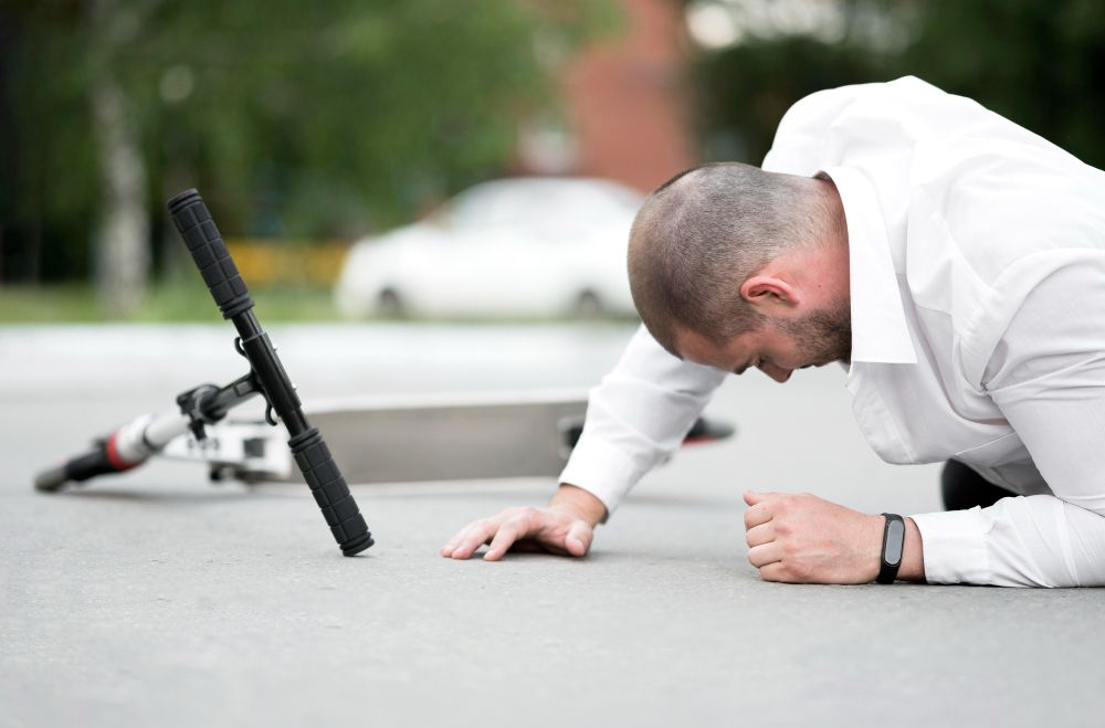 Man on ground next to fallen scooter, possibly after a fall. Asphalt setting, car in the background.