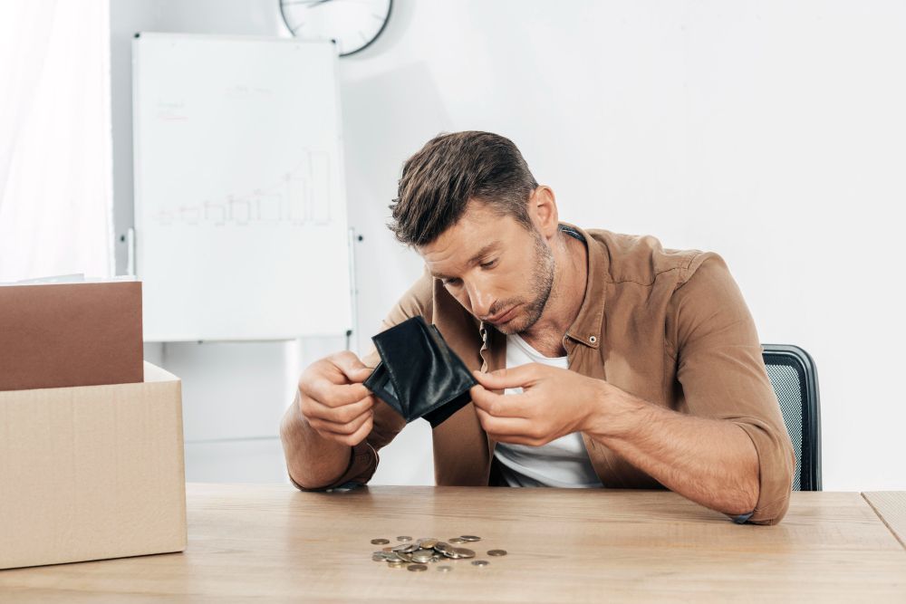 Man looking at empty wallet with few coins on table. Office setting.