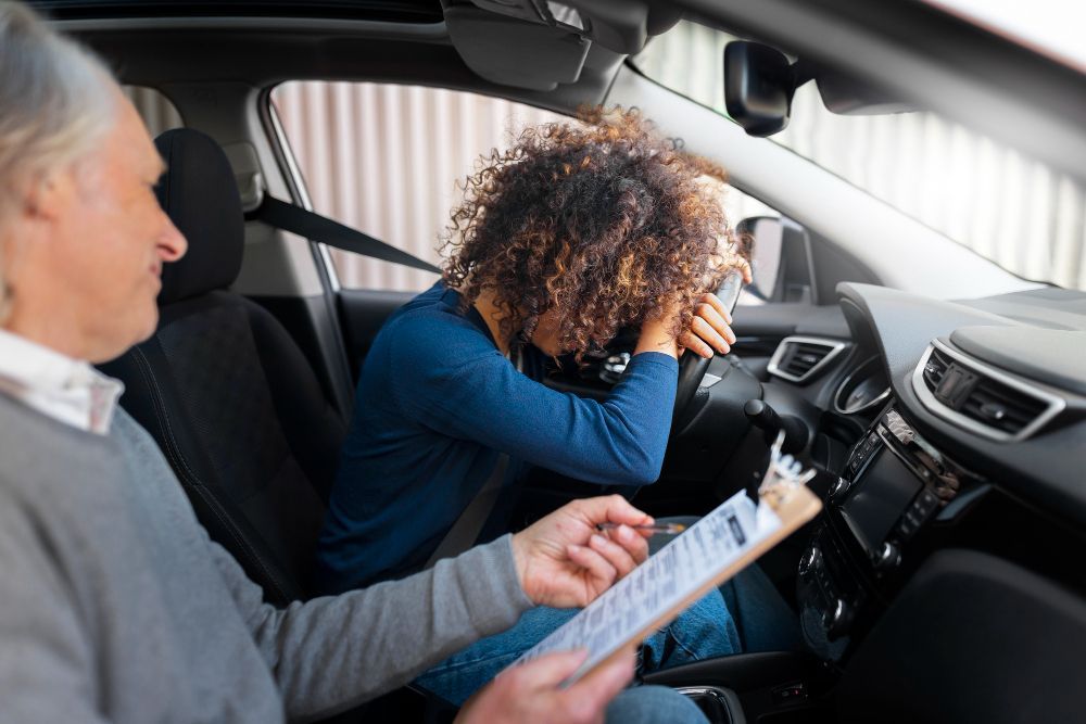 Driving instructor observes student driver, who rests head on steering wheel in apparent distress.