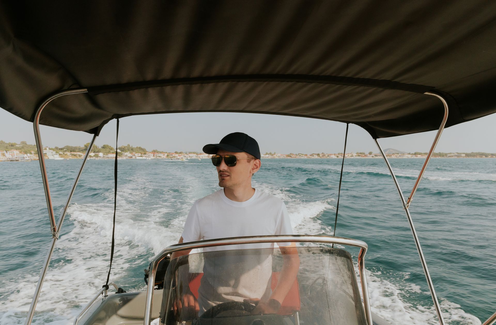 Man wearing sunglasses and a cap, steering a motorboat on a sunny day.
