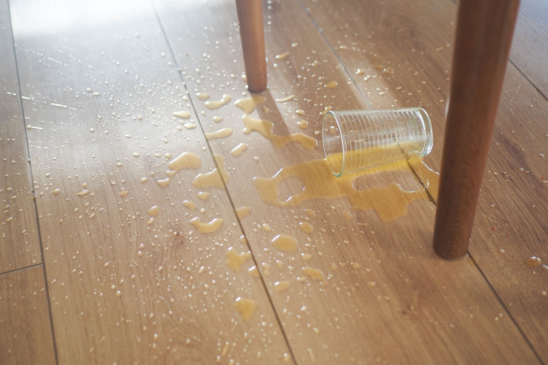 Glass of spilled orange liquid on a wooden floor near a chair leg.