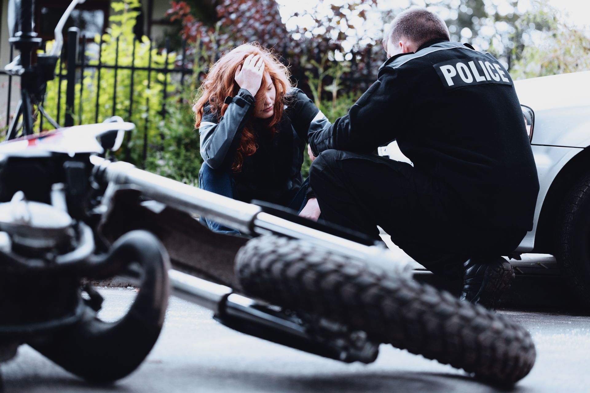 A woman with red hair reacts near a crashed motorcycle as a police officer kneels beside her.