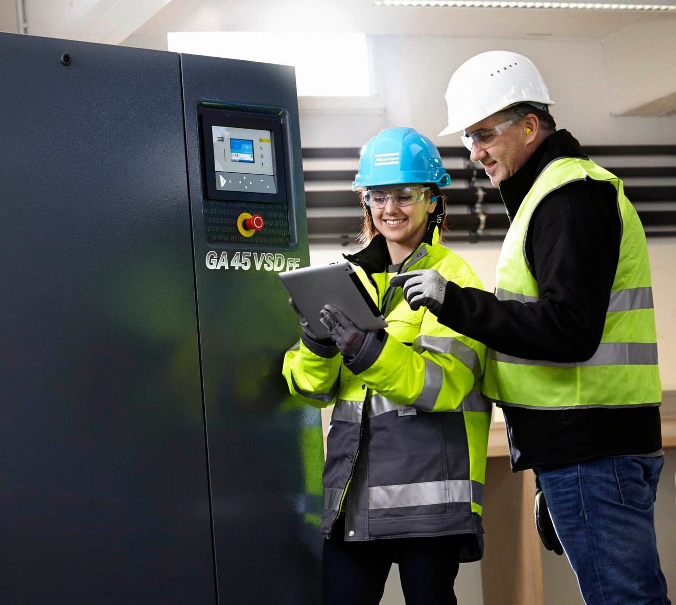 Twee werknemers met veiligheidshelmen bekijken een touchscreen-bedieningspaneel in een industriële omgeving.