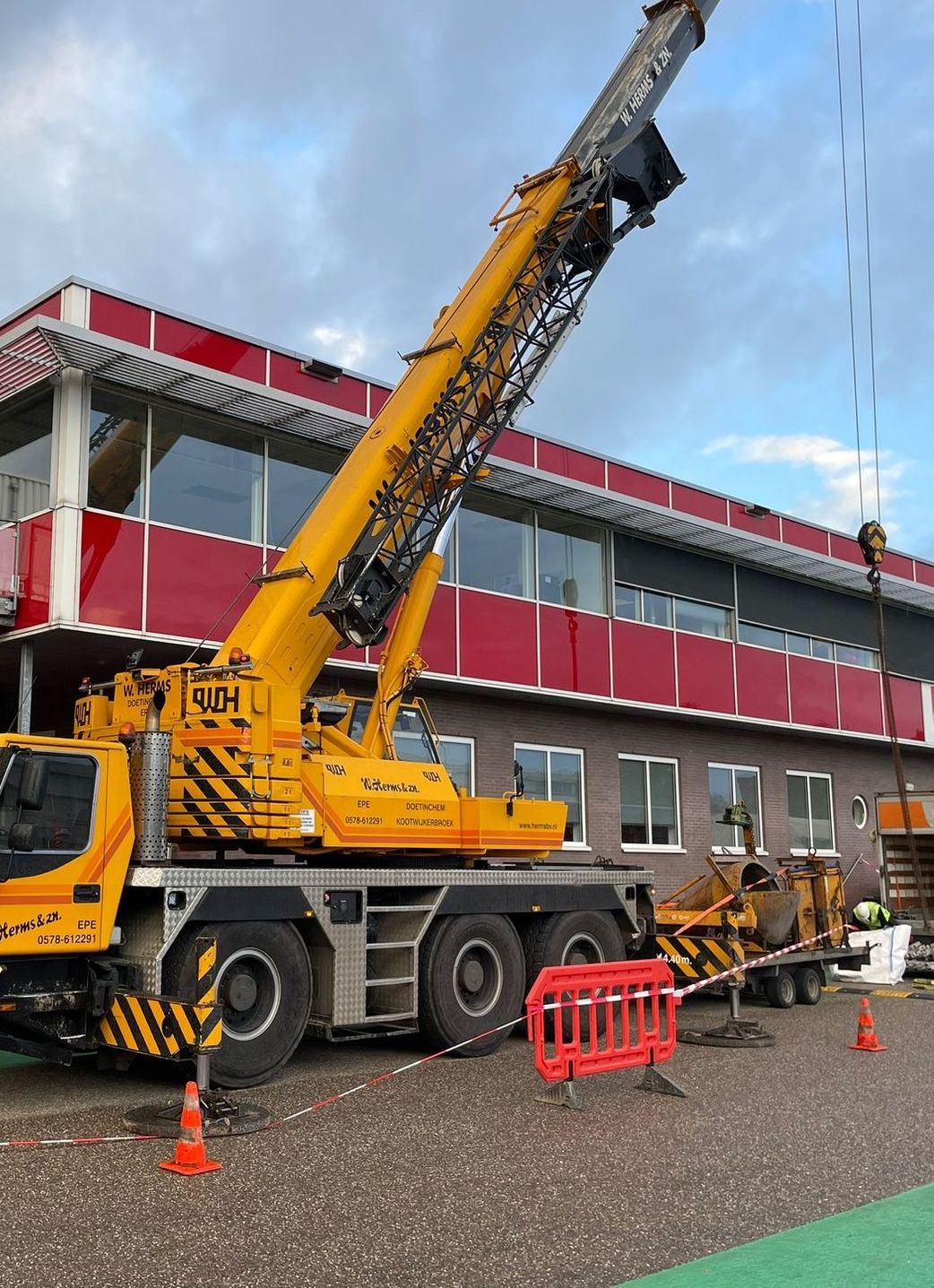 Een gele mobiele kraan staat geparkeerd naast een gebouw, omringd door kegels en afzettingen op een nat terrein.