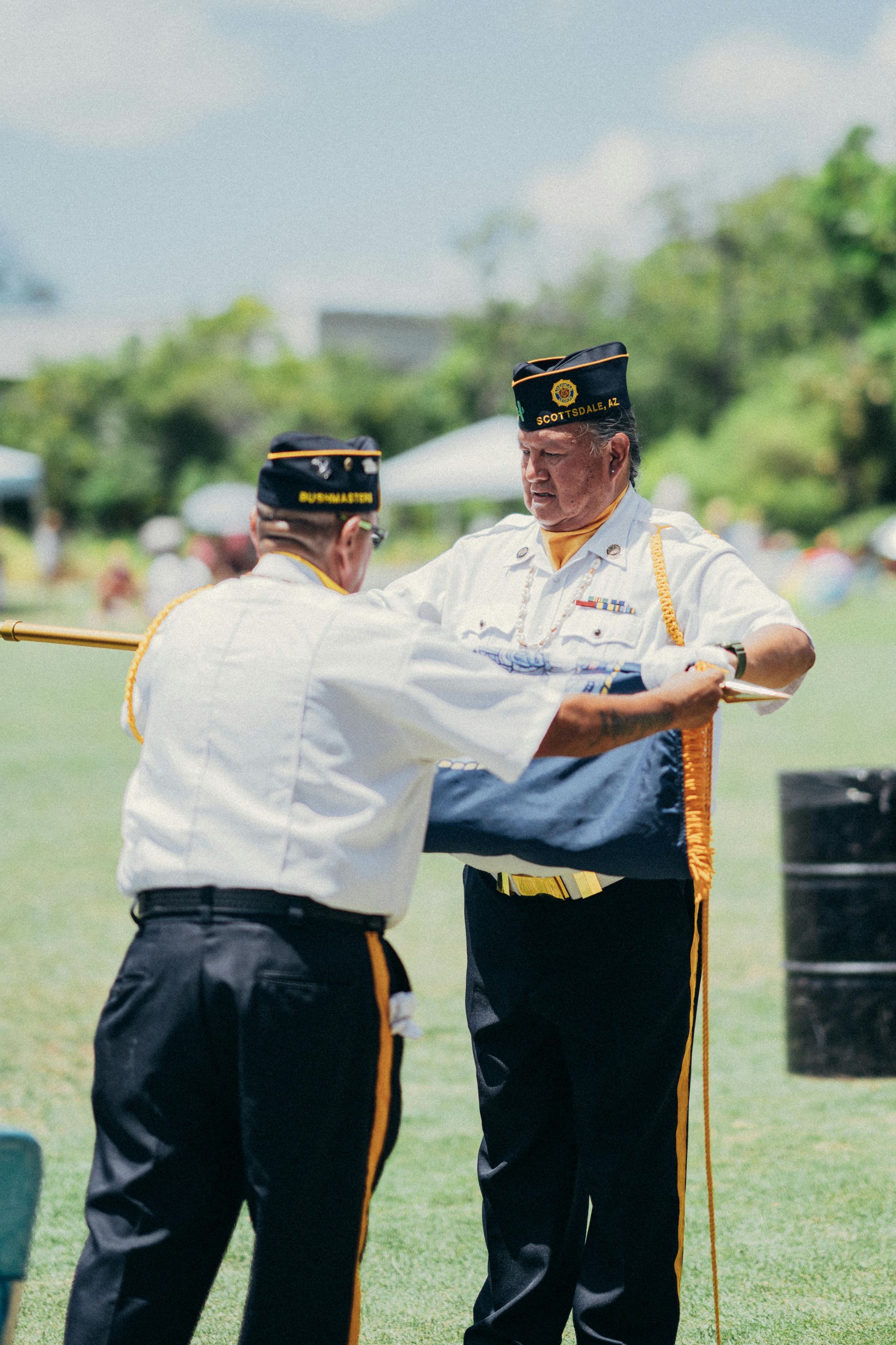 Two men in white uniforms and black hats perform a ceremonial dance on a grassy field.