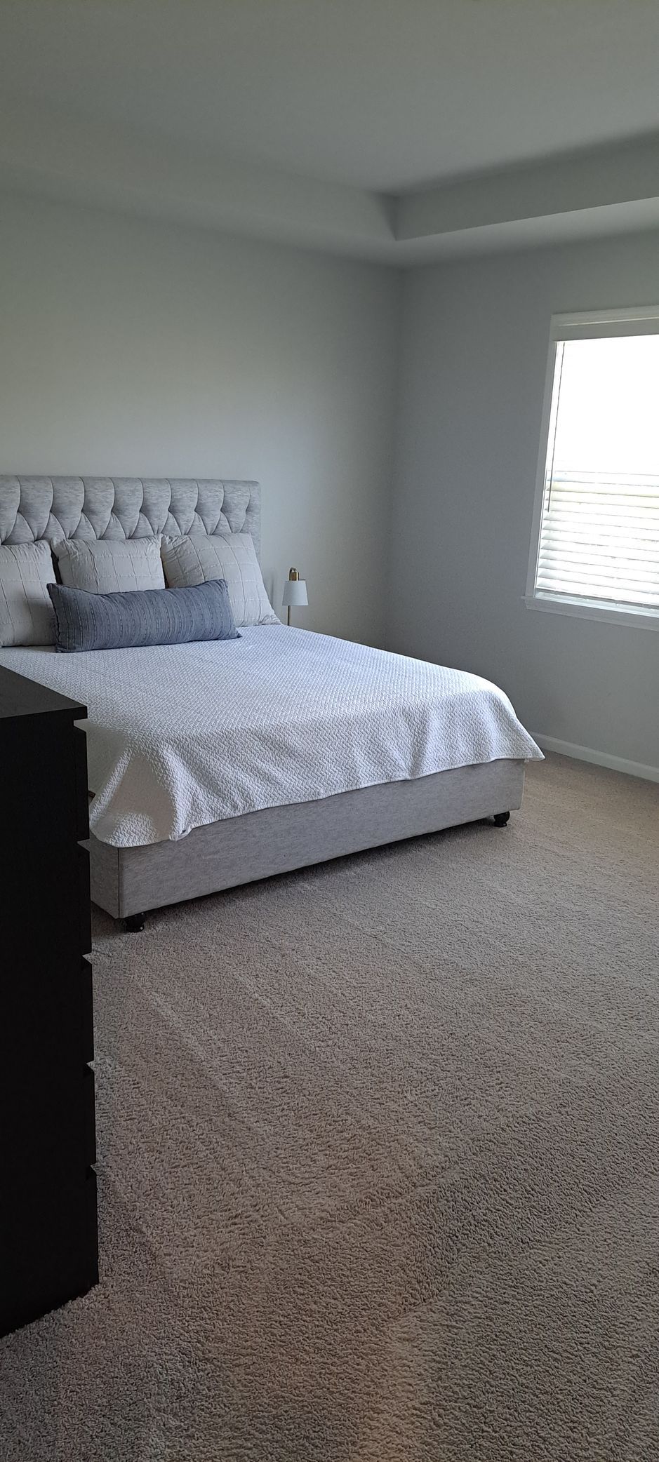 Bedroom with tufted headboard, soft white bedding, and light-colored carpet. Window with blinds.