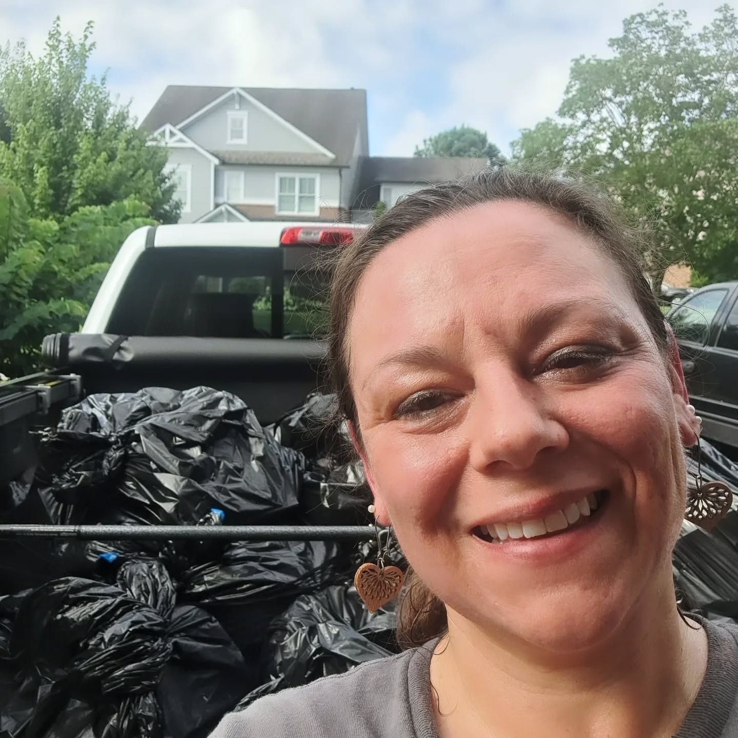 Woman smiling in front of truck bed filled with black trash bags; a house and trees in background.