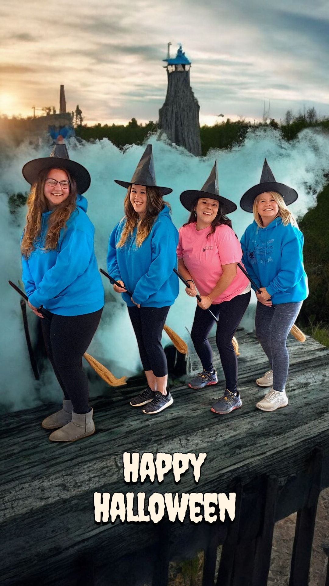 Four people in witch hats and blue/pink tops, holding brooms, standing on a bridge, with smoke and a castle, Halloween message.
