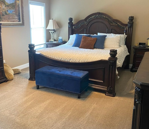 Bedroom with ornate dark wood bed, blue bench, beige carpet, and decorative pillows.