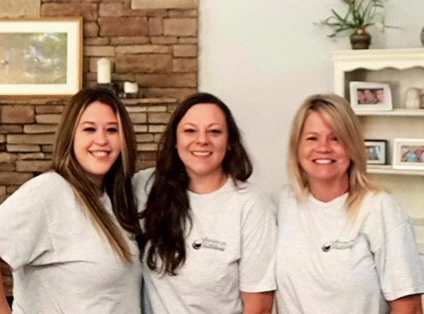 Three women in white shirts smiling, posing in front of a stone wall and a shelf with decor.