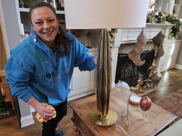 Woman in blue hoodie dusting a tall gold lamp in a living room with Christmas decorations.