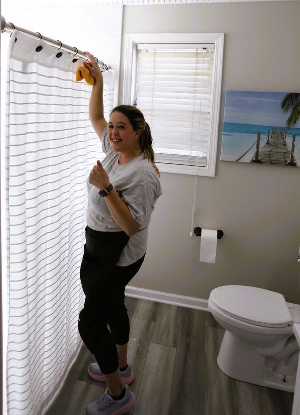 Woman cleaning striped shower curtain in a bathroom with a window and a toilet.