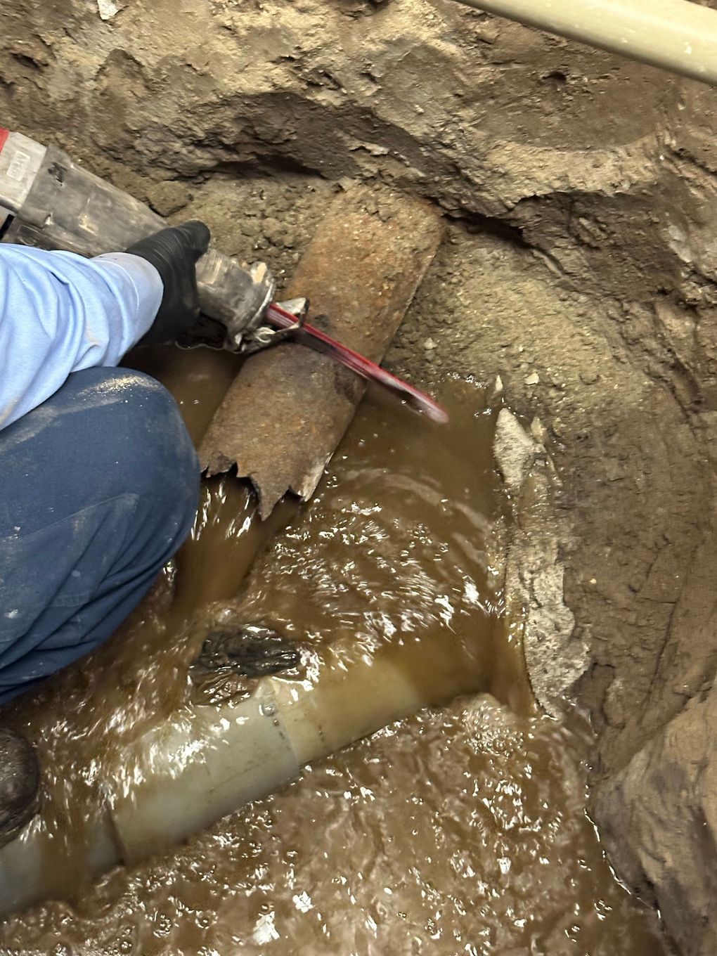A person is kneeling in the dirt next to a broken pipe.