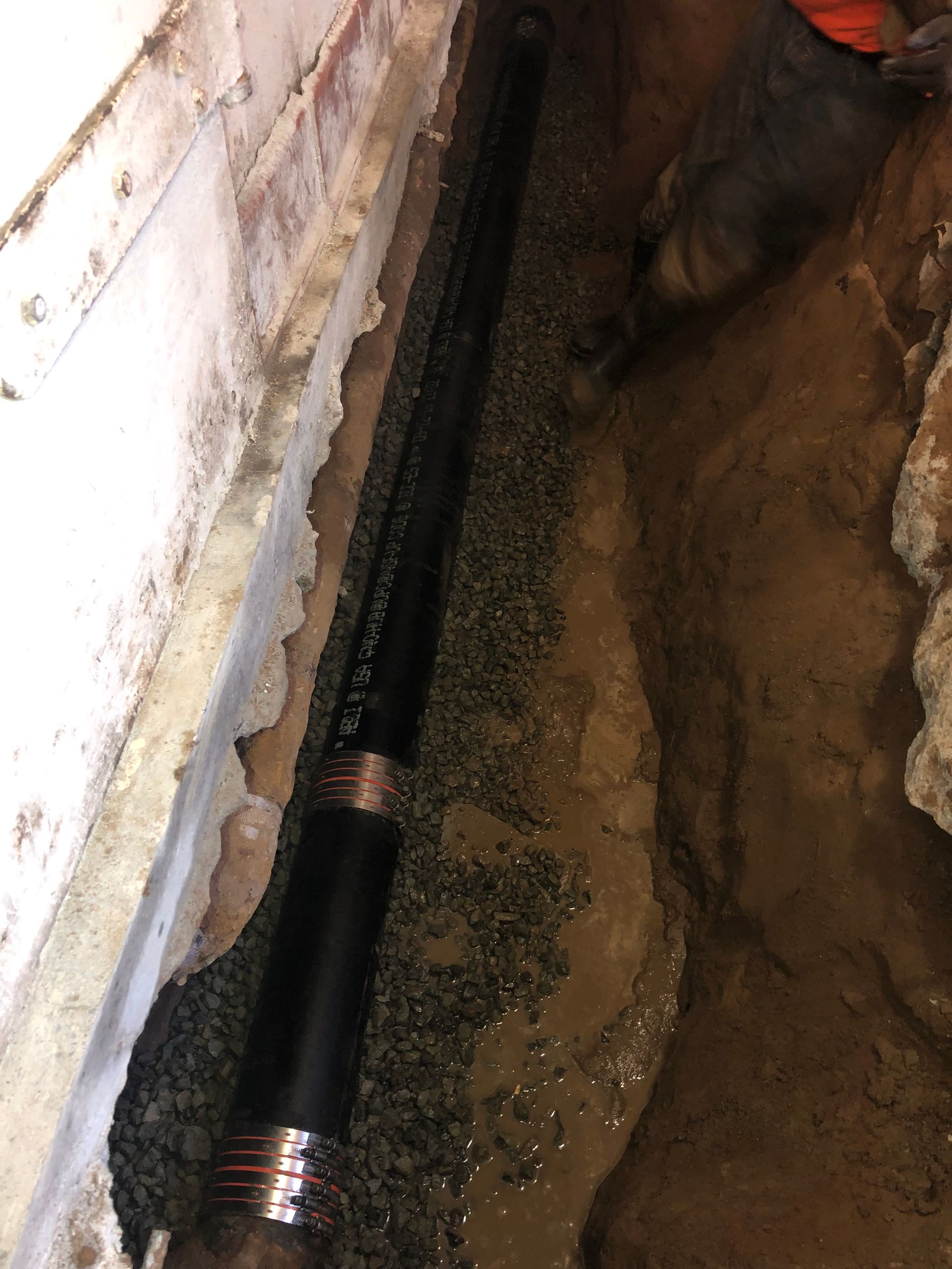 A black pipe is laying in the dirt next to a brick wall.