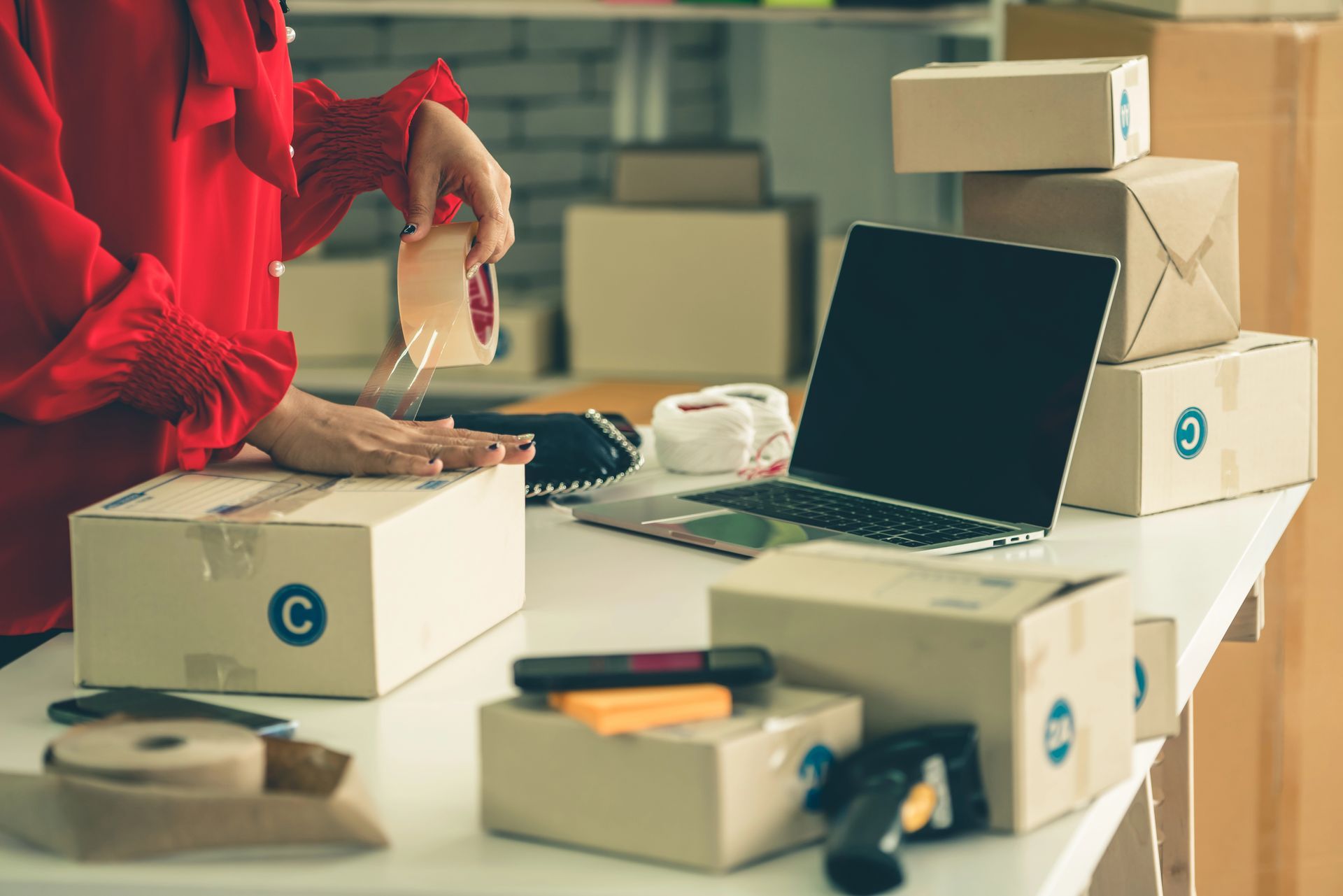 Una persona imballa scatole per spedizioni accanto a un computer portatile aperto su una scrivania disordinata.