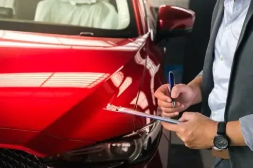 A man is writing on a clipboard in front of a red car.