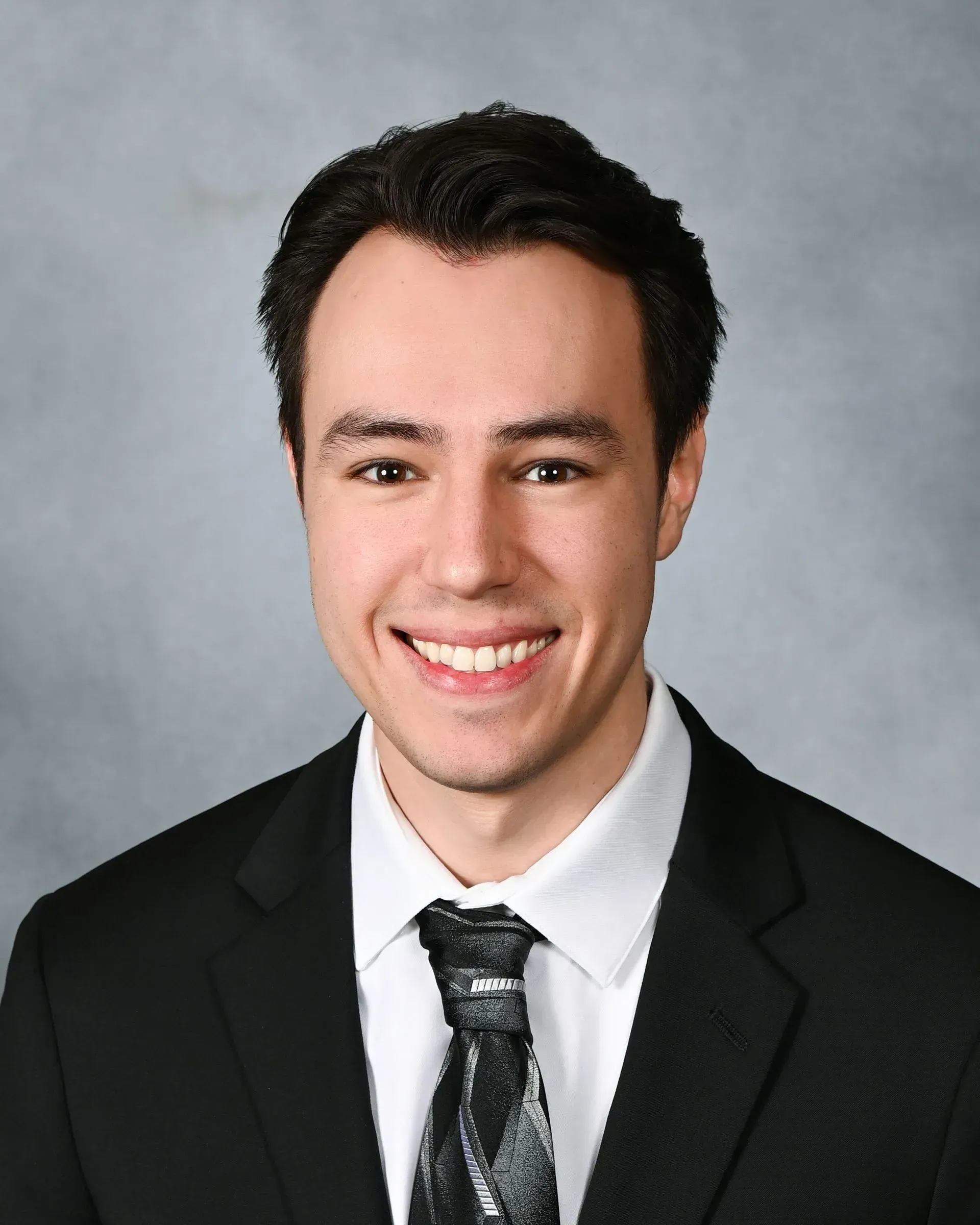 Smiling person in a black suit and tie against a gray studio background