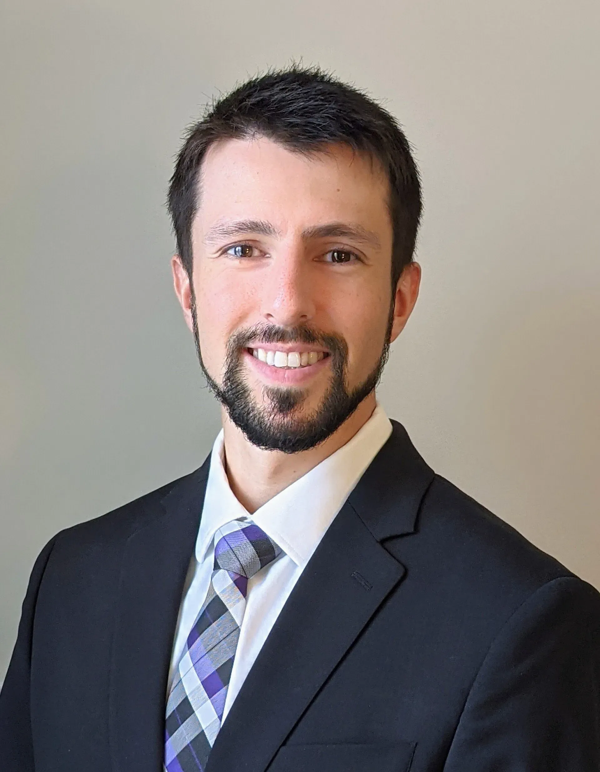 Portrait of a smiling man in a dark suit, white shirt, and patterned tie against a neutral background