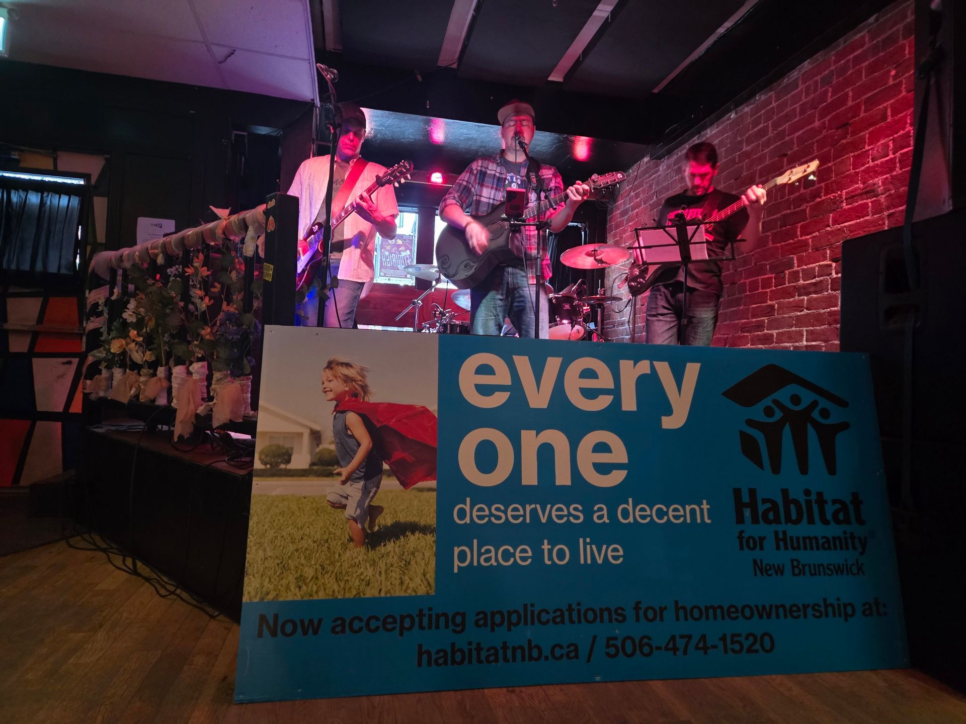 Live band on a small stage behind a Habitat for Humanity New Brunswick banner at a fundraiser event