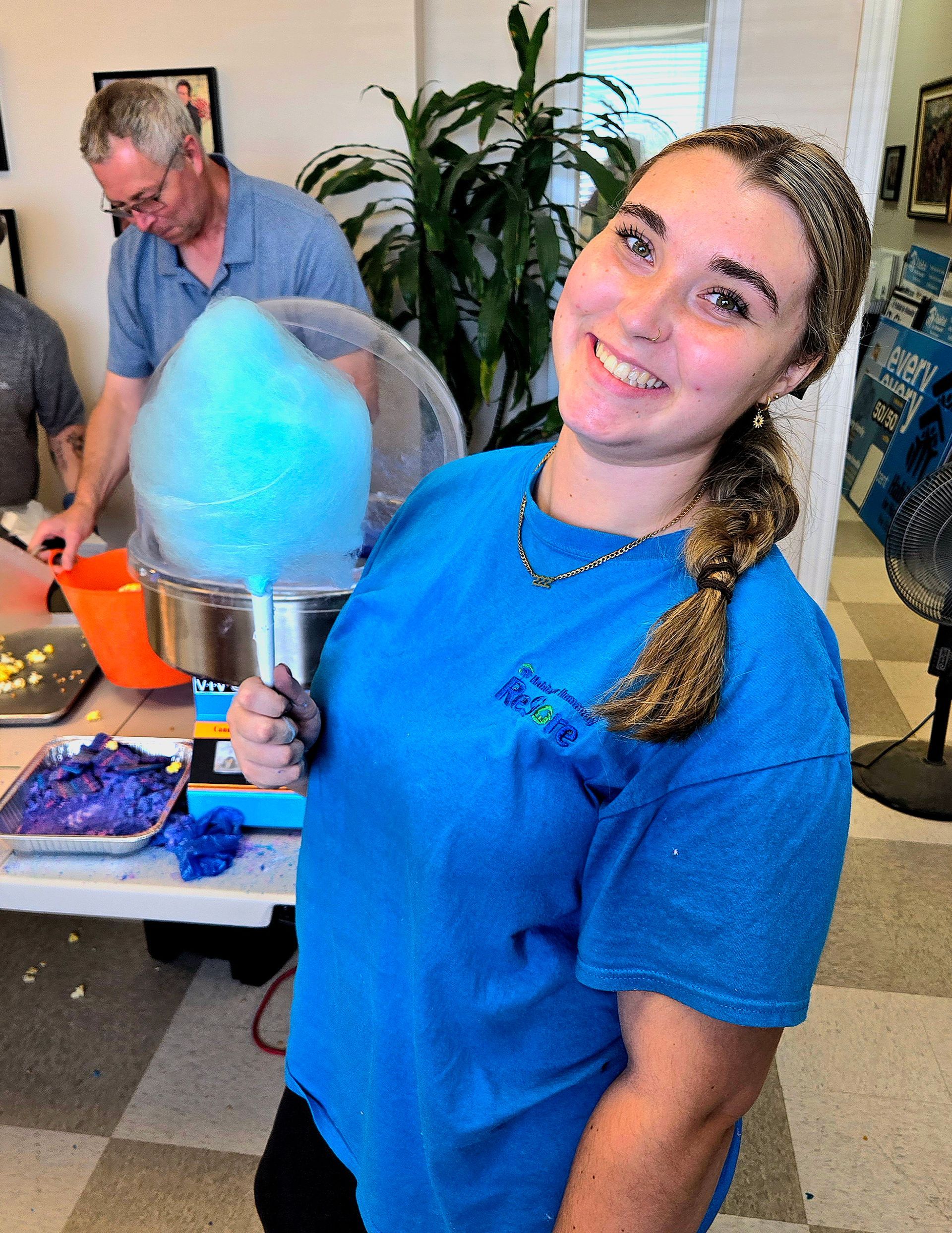 Habitat New Brunswick volunteer in blue shirt holding cotton candy at an indoor fair table, smiling at the camera.