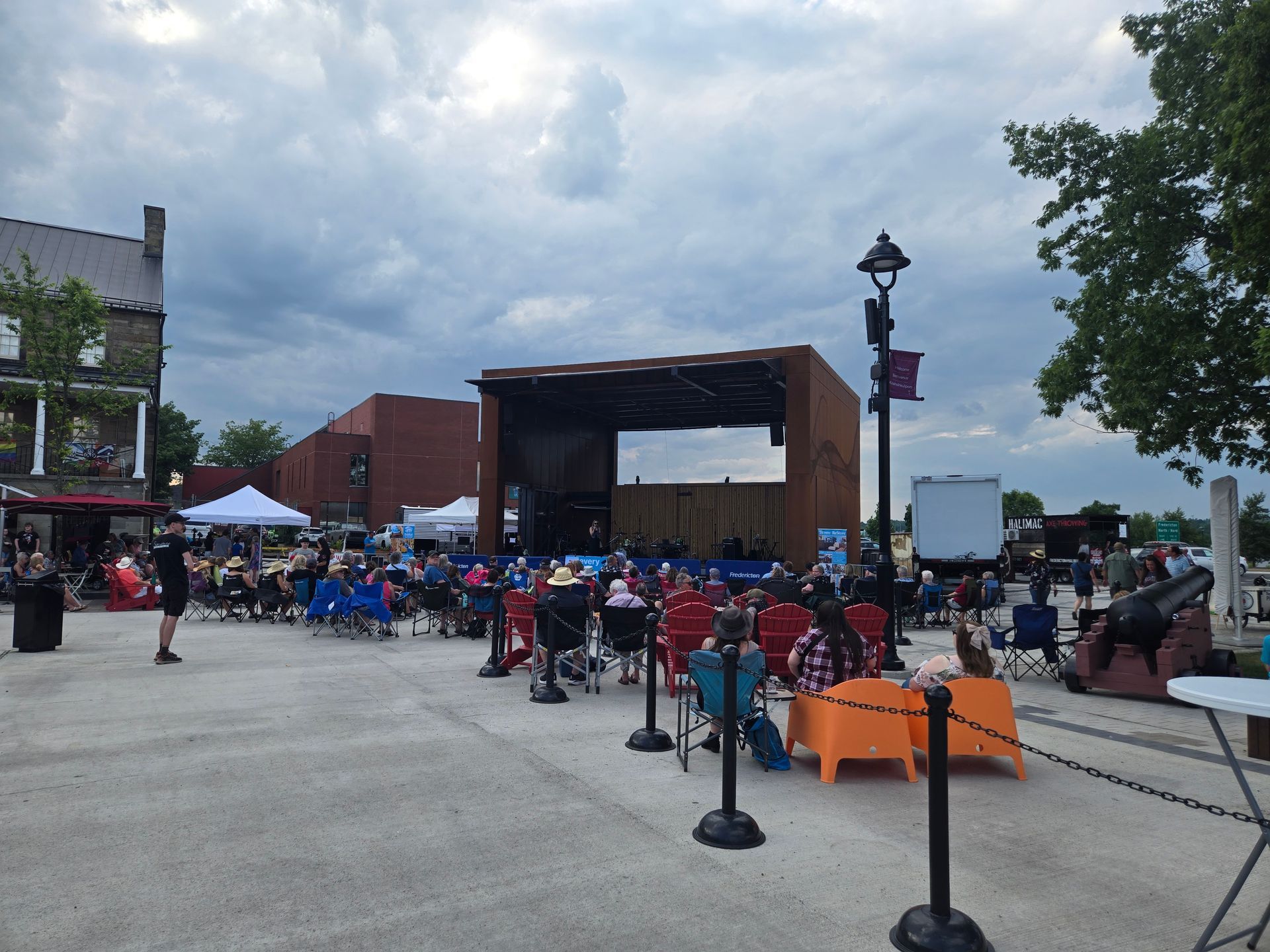 Habitat New Brunswick Outdoor stage in a plaza with seated crowd, tents, and cloudy sky at a community event
