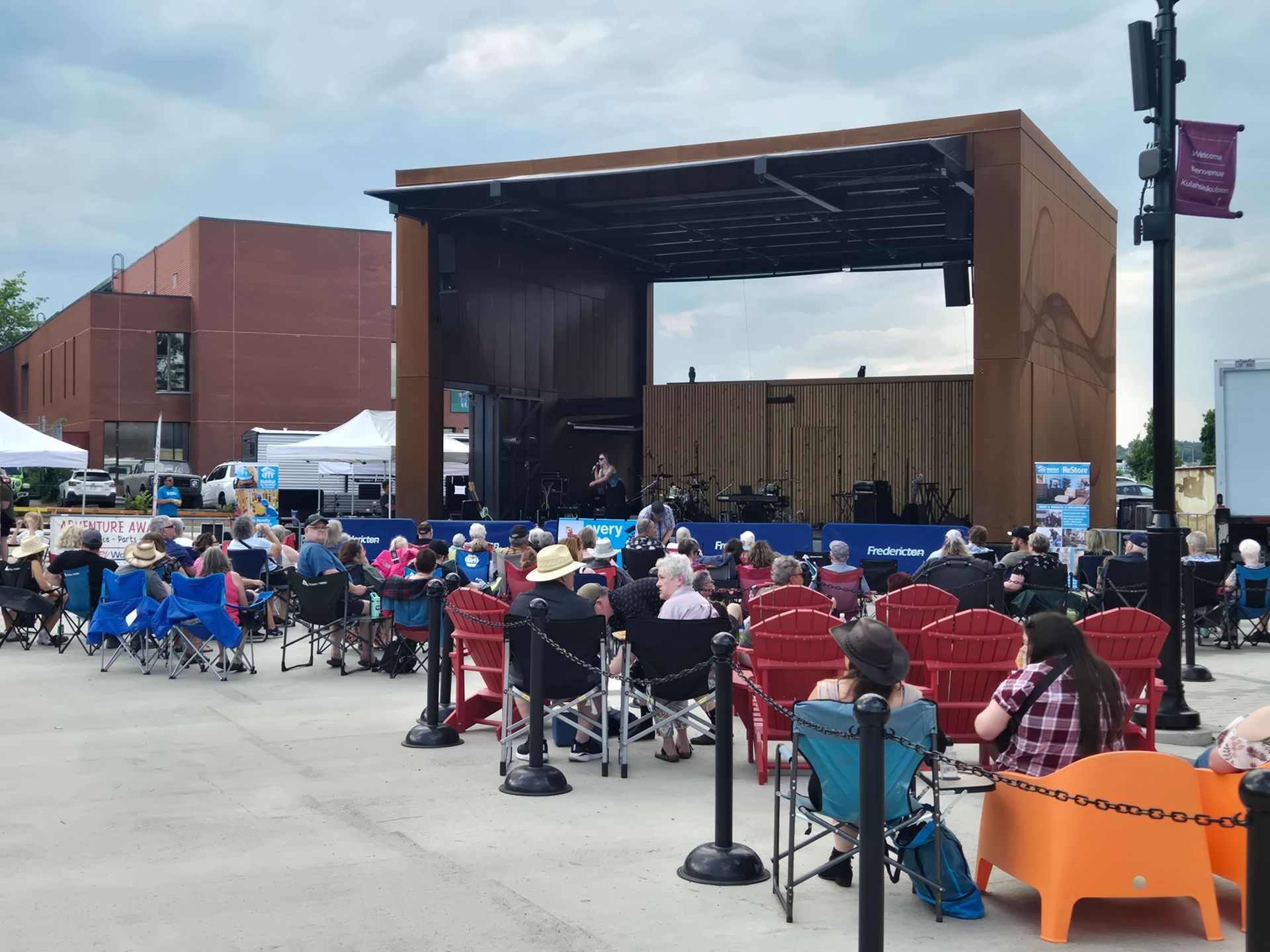 Habitat New Brunswick Outdoor concert audience seated in folding chairs before a large wooden stage in a city plaza