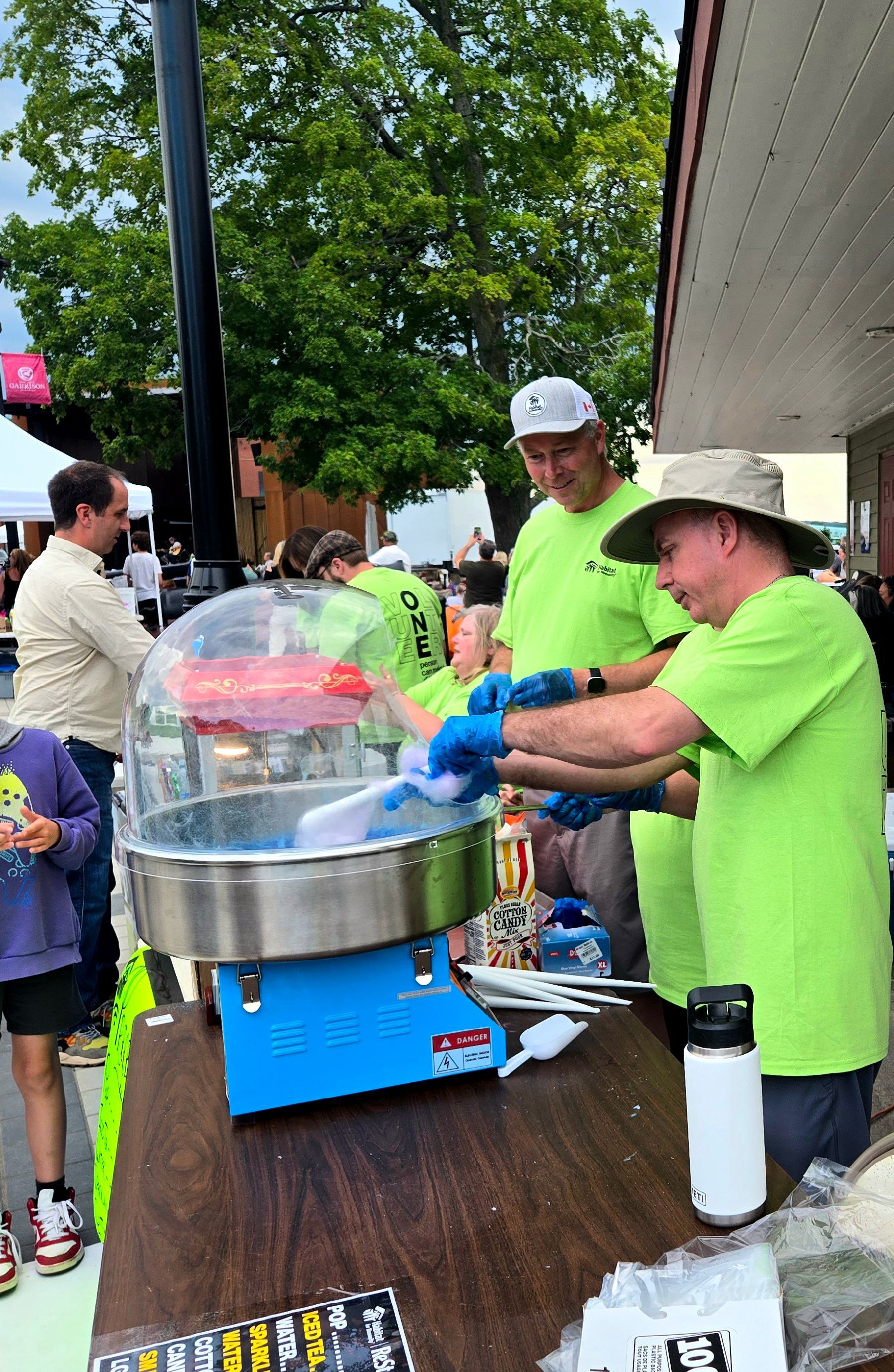 Two Habitat New Brunswick volunteers in neon green shirts serve blue cotton candy at a street fair table