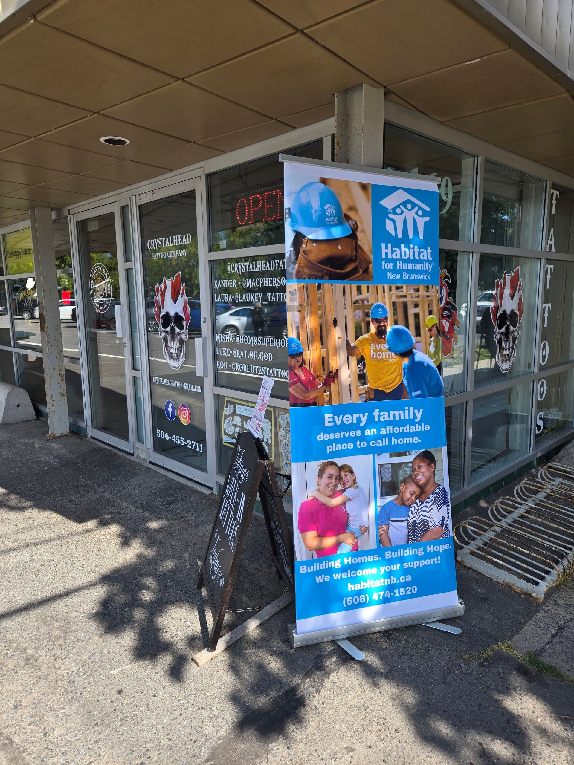 Blue charity banner outside storefront with photos of smiling families and children on the sidewalk, Habitat NB