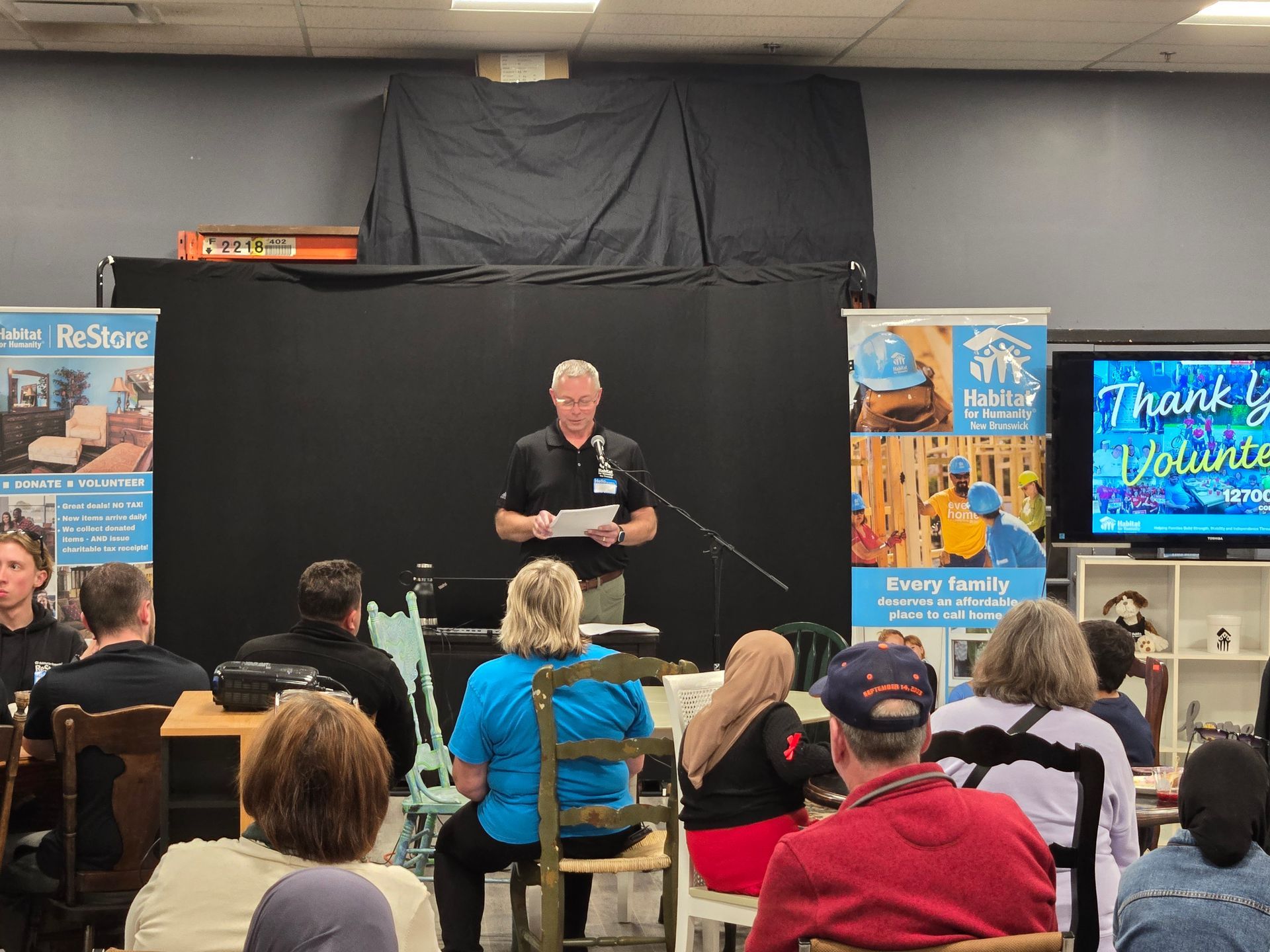 Perry Habitat NB, at a podium addresses seated audience in a community room with banners and posters.