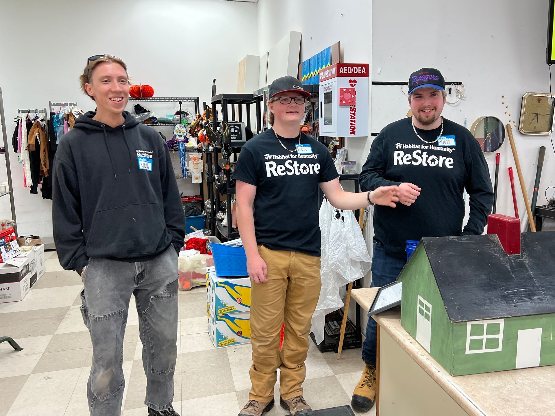 Three Habitat New Brunswick volunteers in ReStore shirts stand beside a small model house in a workshop.