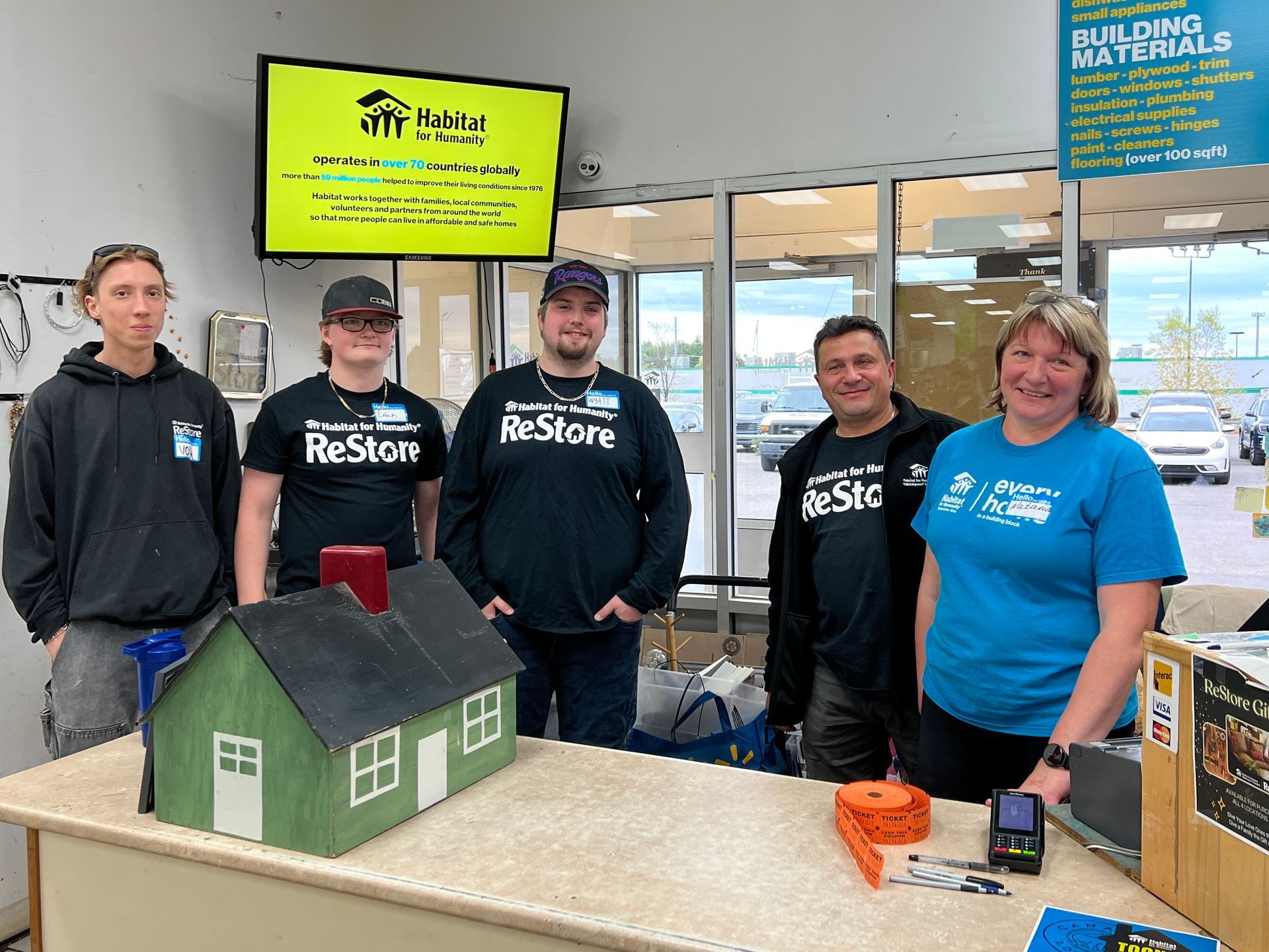 Five Habitat New Brunswick volunteers stand behind a model house display in a bright store near a building materials sign.